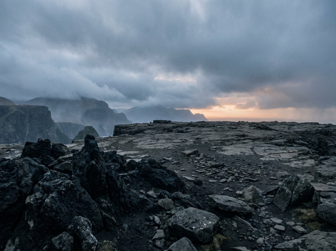 A flat ledge of dark grey stone with black volcanic rocks in front, misty cliffs on the left and a heavy grey sky with a faint orange glow on the horizon.