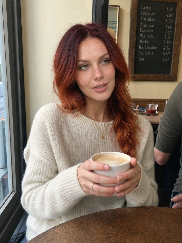Red-haired woman in a cream sweater holding a coffee cup at a café table