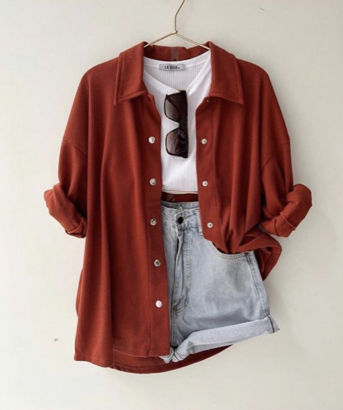 A flat lay of an oversized rust coloured button up shirt worn open over a white ribbed top, with light wash denim shorts and dark sunglasses, hanging on a hanger against a cream wall.